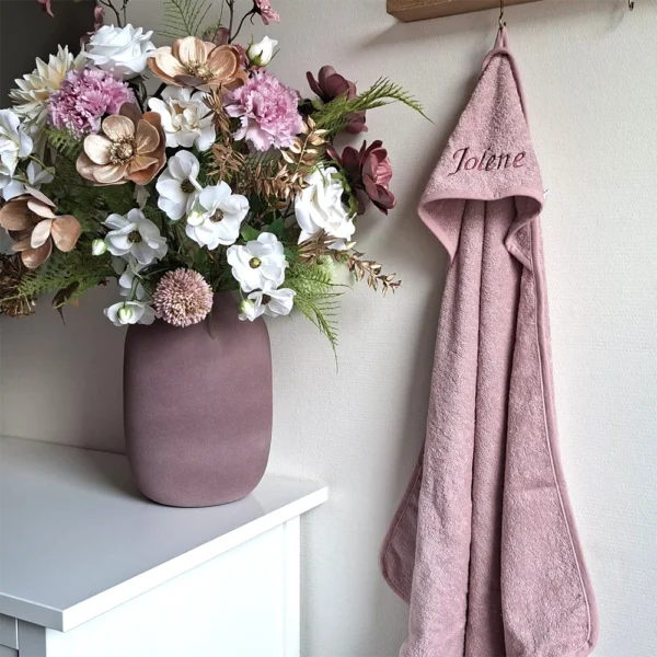 Roze badcape met capuchon, gepersonaliseerd met de naam ‘Jolene’ in sierlijke geborduurde letters, hangend aan een haak naast een decoratief boeket van roze, witte en gouden bloemen.