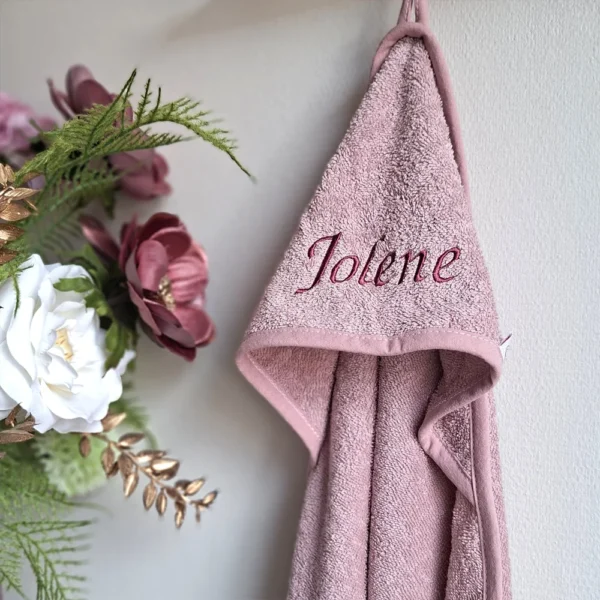 Roze badcape met naam ‘Jolene’, geborduurd met sierljike letters op de capuchon, hangend aan een haak naast een sierlijk boeket met roze, witte en gouden bloemen.