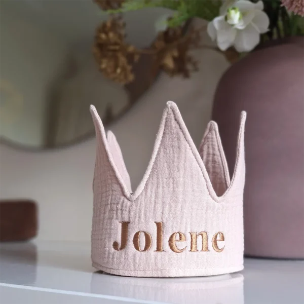 Roze hydrofiele kroon met de naam ‘Jolene’ geborduurd in koperkleurige letters, staand op een witte commode naast een decoratieve vaas met bloemen.