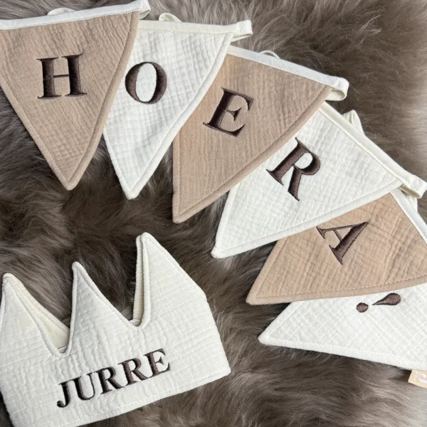 Gepersonaliseerde vlaggenlijn met de tekst “HOERA!” en een bijpassend stoffen kroontje met de naam “Jurre”, beide gemaakt van zachte mousseline stof in neutrale beige en ivoor tinten. Perfect als duurzame feestdecoratie voor verjaardagen of ter decoratie op de kinderkamer