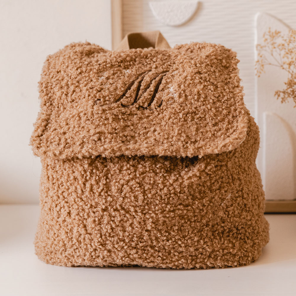 Zachte bruine rugzak van teddystof, gepersonaliseerd met een sierlijke donker bruine letter “M” en kleine witte vlindertjes op de sluiting.