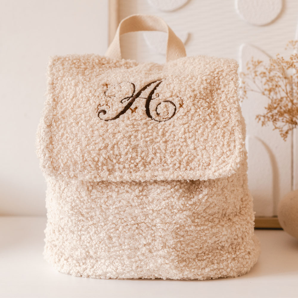 Zachte crème rugzak van teddystof, gepersonaliseerd met een sierlijke bruine letter “A” en kleine koperkleurige vlindertjes op de sluiting.