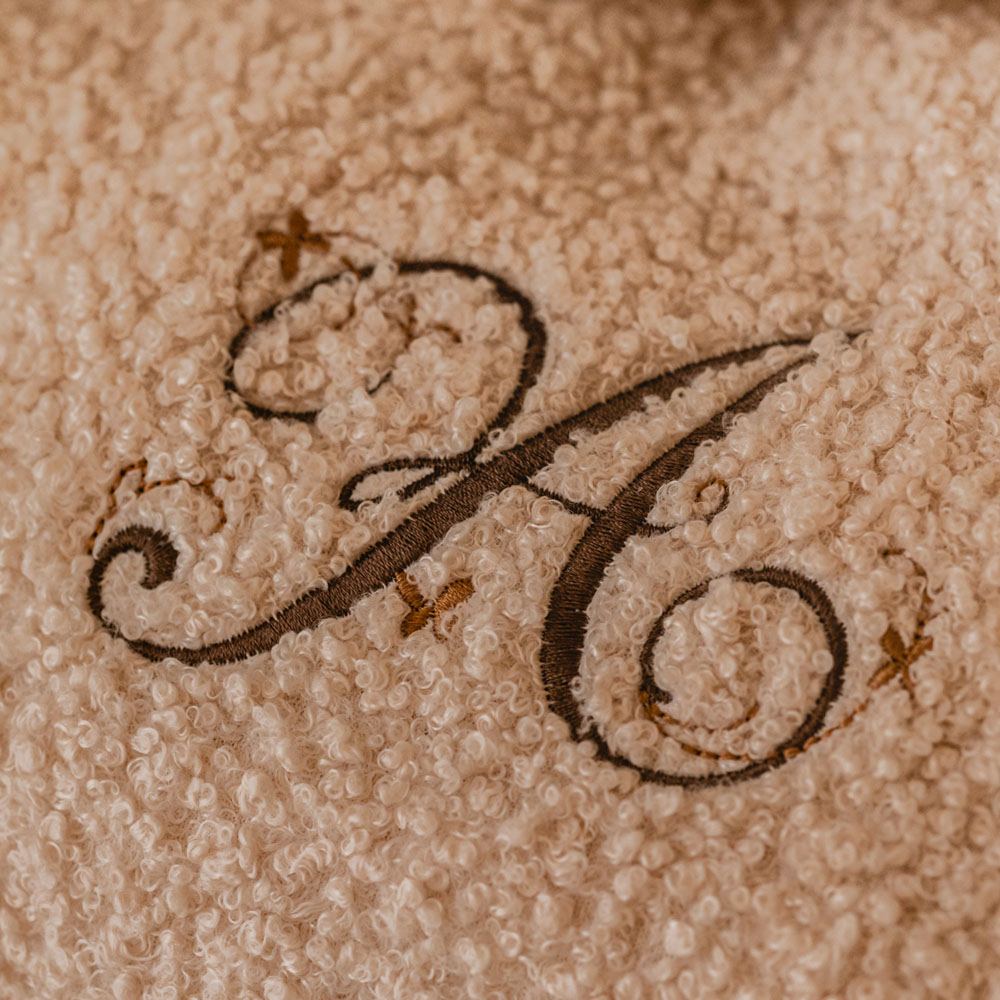 Zachte crème rugzak van teddystof, gepersonaliseerd met een sierlijke bruine letter “A” en kleine koperkleurige vlindertjes op de sluiting.